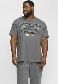 Graues T-Shirt mit kurzen Ärmeln, bedruckt mit "JACK & JONES AUTHENTIC MANUFACTURE" in Gelb und Schwarz, kombiniert mit hellgrauen Jeans.