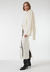 Cremekleurig oversized top met gedrapeerde mouwen, gecombineerd met een aansluitende cremekleurige rok met zwarte en gouden bloemenborduursels en een hoge split. Zwarte hoge laarzen.