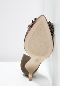 Chaussure à talon aigu en suède brun, à bout pointu et semelle texturée, présentée debout sur un fond blanc en coin.