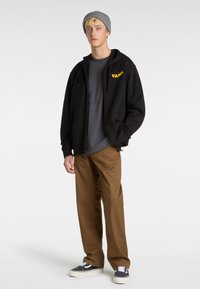 Black zip-up hoodie with yellow "VANS" logo, gray t-shirt, brown pants, and gray beanie, paired with black and white sneakers.
