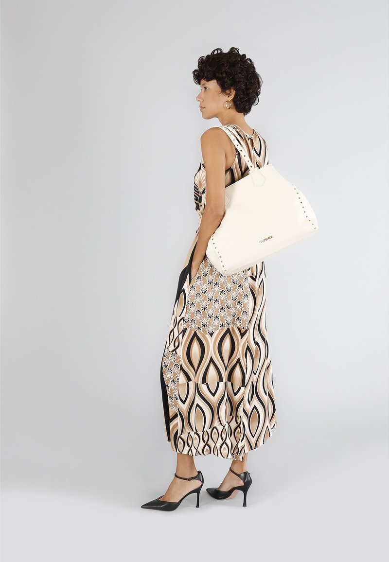 Borsa in pelle bianca con dettagli borchiati, forma strutturata e manico robusto, abbinata a un vestito stampato in tonalità neutre.
