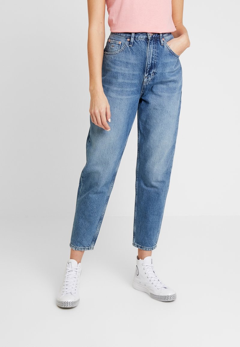 Jean bleu clair taille haute avec une jambe droite et un ourlet retroussé. Associé avec des baskets montantes blanches et un T-shirt rose.