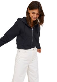 Sweatshirt cropped preto feito de tecido macio, com fecho em zíper na frente, capuz com cordão e bolsos canguru frontais, combinado com calças brancas.