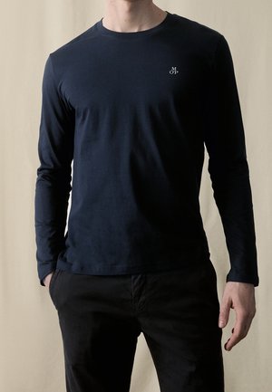 Personne portant un t-shirt à manches longues bleu marine foncé uni avec un petit logo blanc sur la poitrine gauche et un pantalon noir, debout avec une main dans la poche.