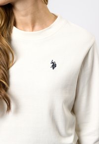 Vrouw die een crèmewitte sweatshirt draagt met een klein donkerblauw geborduurd polo speler logo op de linkerborst.