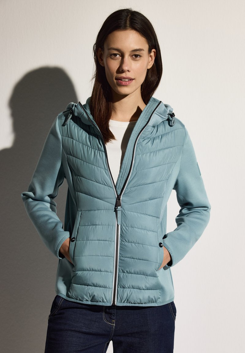 Giacca azzurro chiaro con parte frontale trapuntata, chiusura con zip, cappuccio regolabile e tasche laterali. Realizzata in tessuto morbido con una superficie liscia.