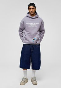 Paarse hoodie met witte tekst, voorzien van verfspattenaccenten. Gecombineerd met losse, donkerblauwe denim shorts en grijze sneakers. Witte sokken zichtbaar.