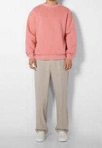 Person som bär en lös rosa sweatshirt, beige vida byxor och vita sneakers, som står mot en helt vit bakgrund.