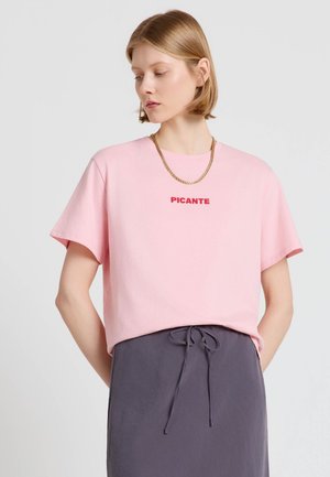 Donna con capelli biondi corti che indossa una T-shirt rosa con la scritta "PICANTE" e una catena d'oro, abbinata a una gonna grigio scuro con coulisse.