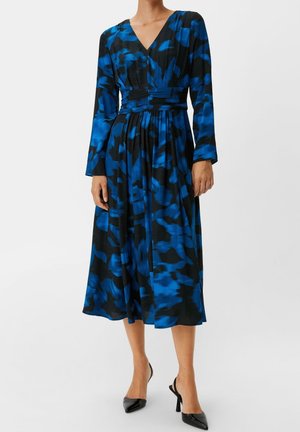 Femme portant une robe midi à imprimé abstrait noir et bleu avec manches longues, col en V, taille cintrée, et des escarpins noirs à bouts pointus.