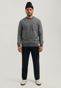 Hombre con un suéter de punto gris con bolsillo en el pecho, pantalones negros, zapatillas blancas y gorro de punto negro, de pie contra un fondo liso.