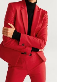 Blazer rouge sur mesure avec boutons noirs, associé à un col roulé noir. Tissu lisse, revers nets et coupe structurée.