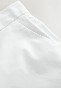 Gros plan sur un tissu blanc montrant une ceinture et un détail de couture sur une surface lisse, légèrement texturée.