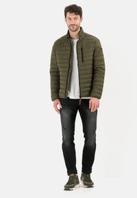 Olivengrøn pufferjakke med høj krave, lynlåslomme og quiltet design, parret med mørke jeans og grønne sneakers.