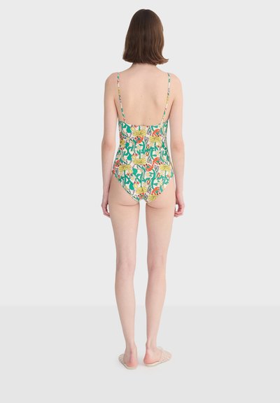 Femme debout de dos, portant un maillot de bain une pièce à motifs floraux avec des bretelles fines et des sandales à enfiler, sur un fond uni.