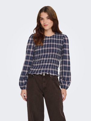 Jeune femme aux cheveux bruns lâchés portant un chemisier smocké à manches longues bleu marine et blanc à carreaux et un pantalon sombre taille haute, debout devant un fond blanc.