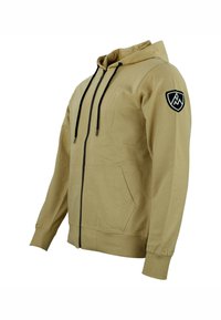 Sudadera con cremallera de un beige claro, con cremallera y cordones negros. Cuenta con bolsillos frontales y un logo bordado en negro en la manga izquierda.