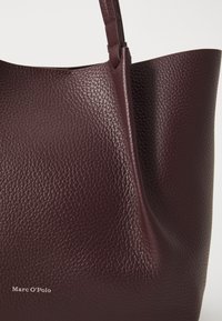 Sac fourre-tout en cuir bordeaux avec finition texturée, présentant une silhouette douce et une seule bandoulière. Logo de la marque Marc O'Polo visible.