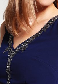 Mörkblå klänning med V-ringning, prydd med intrikata pärlbroderier längs halsringningen i silver och mörka nyanser, som framhäver ett strukturerat tyg.