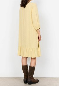 Robe jaune à motifs avec un ourlet volanté, des manches trois-quarts et une coupe décontractée, associée à des bottes brunes jusqu'au genou.