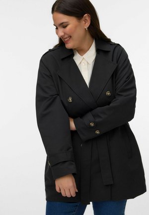 Femme souriante regardant vers sa gauche, portant un trench-coat noir ceinturé sur une chemise blanche à col et un jean bleu, les bras croisés devant elle.