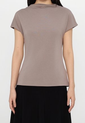 Kvinde iført en taupe kortærmet top med en draperet halsudskæring og et sort skørt, der står foran en ensfarvet hvid baggrund.