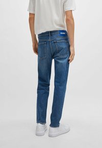 Blue denim jeans with a straight cut, featuring back pockets and a branded waistband. Paired with white sneakers.