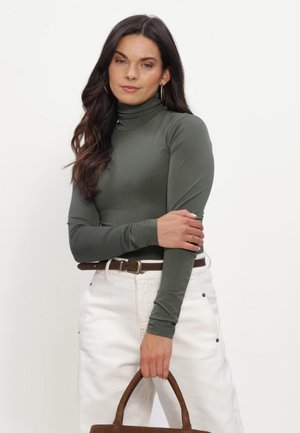 Vrouw met donker haar draagt een groene coltrui met lange mouwen, witte broek met een bruine riem en houdt een bruine handtas vast voor een effen achtergrond.