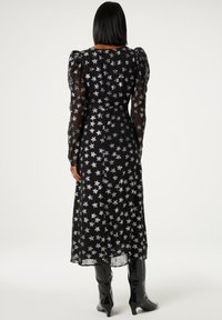 Schwarzes Kleid mit durchsichtigen langen Ärmeln, verziert mit silbernen Sternmustern, mit tailliertem Schnitt und fließendem Rock, kombiniert mit kniehohen schwarzen Stiefeln.