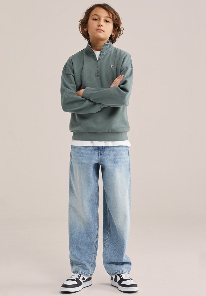 Laps seisab käed risti, seljas roheline poollukuga dressipluus, helesinised laiade säärega teksad ja mustvalged tossud.