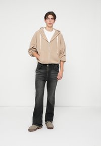 Corta-vento bege com fecho éclair e cordões, camiseta branca por baixo, jeans pretos à boca-de-sino com detalhes desgastados e botas cinzentas de tornozelo.