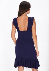 Marineblauw gebreid jurk met ribbelstructuur, volants mouwen en een lage rug. De zoom heeft een uitlopende volants ontwerp.