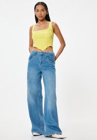 Top cortado amarillo con tela texturizada y escote cuadrado, combinado con jeans de pierna ancha azul claro, que cuentan con bolsillos delanteros y cierre de botón.
