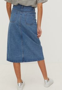 Kvinna som bär en blå denim midi kjol, randig skjorta instoppad, och vita sneakers med silverklackar, sedd bakifrån.