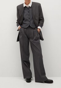 Szary garnitur na miarę z długim żakietem, kamizelką na guziki i szerokimi spodniami. Wykonany z teksturowanej tkaniny, pod spodem biała koszula z kołnierzykiem.