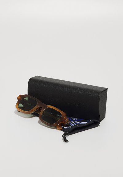 Ochelari de soare din plastic maro, cu lentile întunecate, având o formă pătrată. Vine cu un toc texturat negru și o cârpă cu un model albastru.