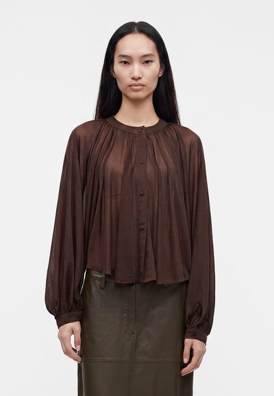 Femme aux longs cheveux noirs portant un chemisier ample plissé marron foncé et une jupe en cuir foncée sur un fond uni.