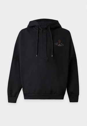 Mand iført sort hættetrøje med lille logo, løse sorte bukser med røde lodrette striber og sorte sko, stående med den ene hånd i lommen.