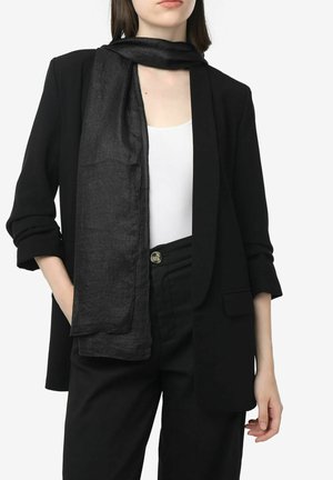 Blazer noir taillé avec des manches retroussées, associé à une écharpe noire transparente sur un haut blanc et un pantalon noir taille haute.