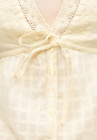 Ρούχο σε κρεμ απόχρωση με δαντέλα πλεξίματος, μπροστινό άνοιγμα με κουμπιά και δεμένο φιόγκο στον λαιμό.