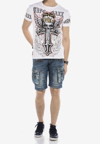 Hombre vistiendo una camiseta blanca con un gráfico de calavera, corona, alas y cruz, pantalones cortos de trabajo azules, zapatillas negras y una pulsera con tachuelas.