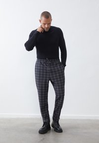Pull noir à côtes, pantalon à carreaux gris avec taille élastique et cordon de serrage, associé à des bottes noires à lacets sur un fond neutre.