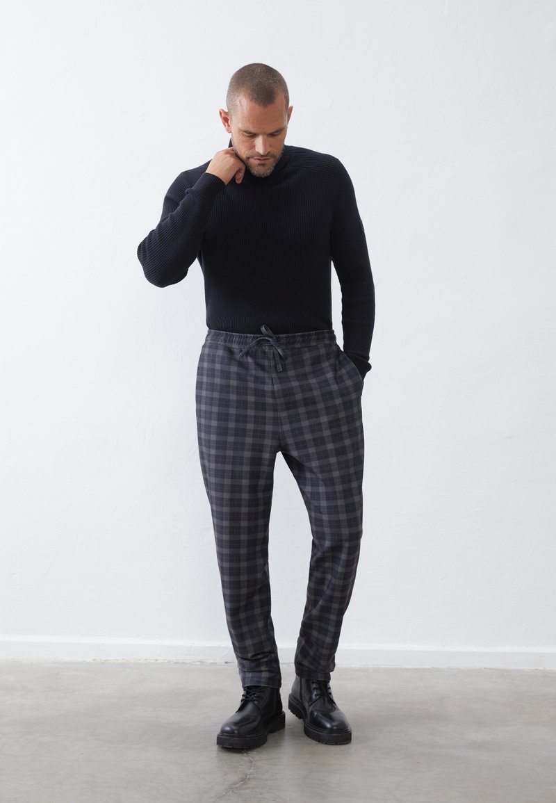 Pull noir à côtes, pantalon à carreaux gris avec taille élastique et cordon de serrage, associé à des bottes noires à lacets sur un fond neutre.