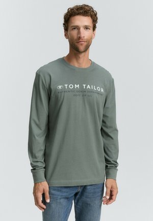 Langärmliges, Rundhals-Shirt aus einem gedämpften grünen Baumwollstoff mit weißem Schriftzug "TOM TAILOR" und Details zur Denim-Abteilung; kombiniert mit blauen Jeans.