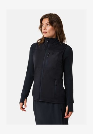 Donna che indossa un gilet nero senza maniche con cerniera sopra una camicia a maniche lunghe, guardando leggermente a sinistra su uno sfondo semplice.