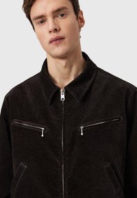 Μαύρο τζάκετ από κορντόρι με φερμουάρ στο μπροστινό μέρος, δύο τσέπες στο στήθος με φερμουάρ και μυτερό γιακά. Το ύφασμα παρουσιάζει διακριτική υφή.