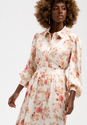 Kobieta z kręconymi włosami ubrana w kwiecistą sukienkę z długim rękawem, zapinaną na guziki, z dopasowaną talią i wzorami w kolorach różowym, kremowym i zielonym.