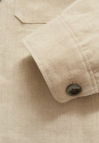 Beige linned skjorteærme med rund kant, udsmykket med en mørk knap med to huller. Subtil tekstur synlig i stoffet.