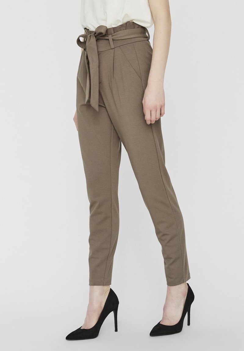 Bruine broek met hoge taille, een strikceintuur, taps toelopende pijpen en plooien aan de voorkant, gecombineerd met zwarte hoge hakken. Glad, rekbaar stoftextuur.