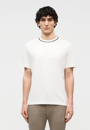 Νεαρός με σγουρά σκούρα μαλλιά, φορώντας απλό λευκό κοντομάνικο μπλουζάκι και μπεζ παντελόνι, στέκεται μπροστά σε λευκό φόντο.